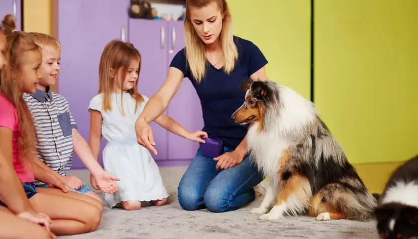 Dzieci i pies uczestniczą w sesji dogoterapii. Kobieta instruuje psa, a dzieci z uwagą obserwują.
