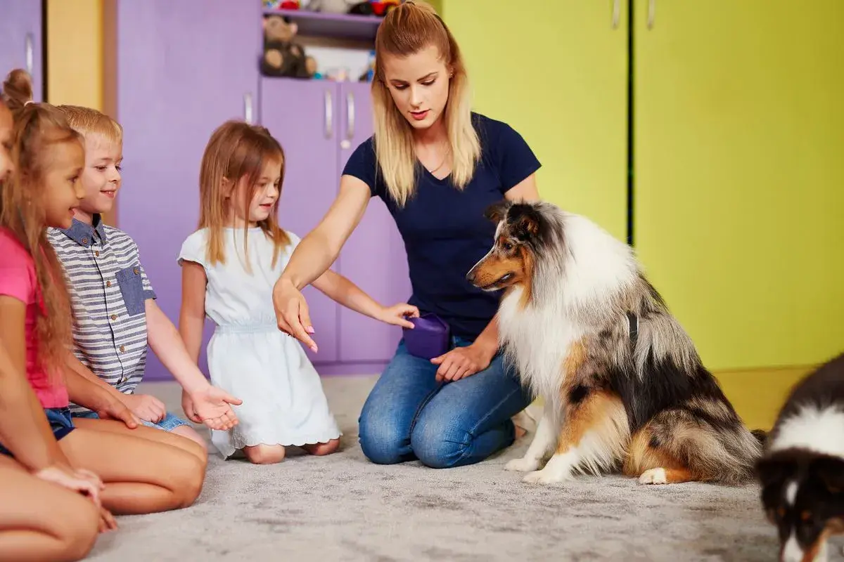 Dzieci i pies uczestniczą w sesji dogoterapii. Kobieta instruuje psa, a dzieci z uwagą obserwują.